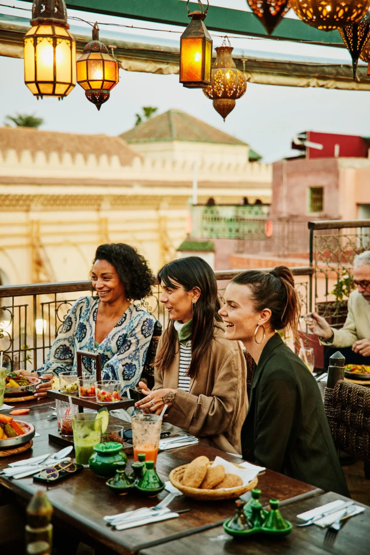 Tres amigas riendo durante una cena en un rooftop iluminado con farolillos