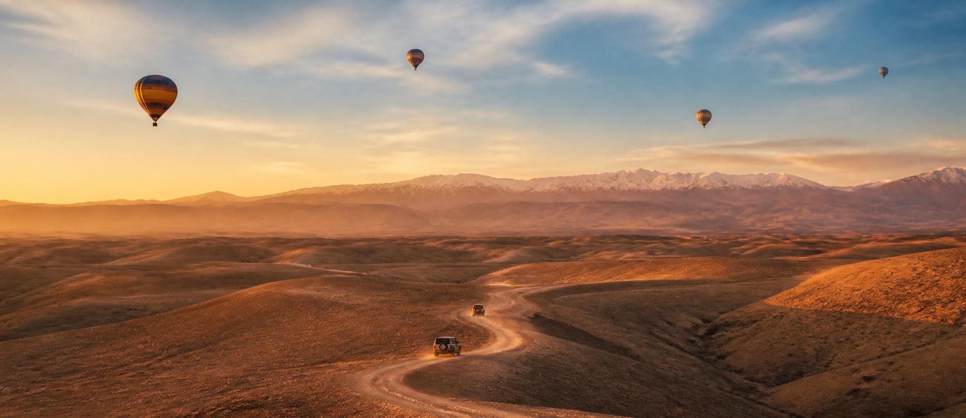 Paysage du désert d'Agafay au lever du soleil, montgolfières dans le ciel, 4x4 sur une piste et Atlas en arrière-plan